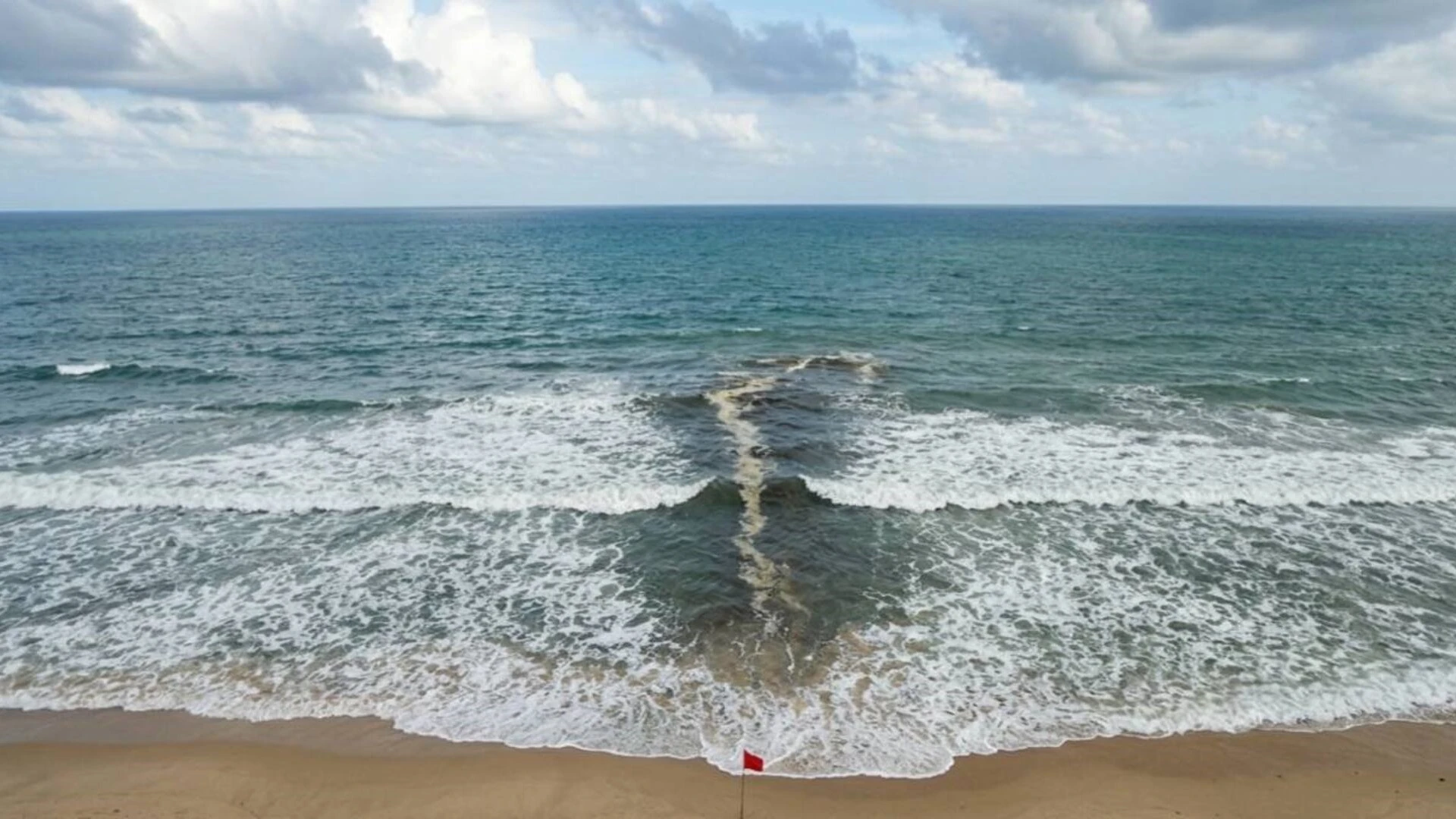A rip current on a Phuket beach seen from above — a narrow dark channel of water cutting through the surf line
